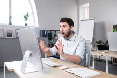 Sinirli, duygusal, sinirli genç bir adam, ofis çalışanı, bilgisayar başında video görüşmesinde konuşan bir yönetici, ofiste beyaz elbiseli bir masada oturuyor.