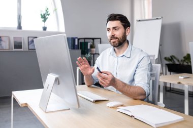 Genç bir adam, bir ofis çalışanı bilgisayarın ofisindeki bir masada oturuyor, bilgisayar kamerasıyla video görüşmesi yapıyor, ortaklarla, müşterilerle iş görüşmesi yapıyor..