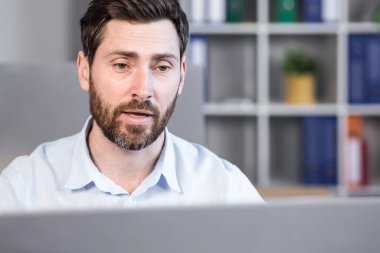 Genç bir adam, bir ofis çalışanı bilgisayarın ofisindeki bir masada oturuyor, bilgisayar kamerasıyla video görüşmesi yapıyor, ortaklarla, müşterilerle iş görüşmesi yapıyor..