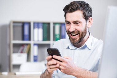 Fotoğrafı kapat. Mutlu ve neşeli bir ofis çalışanı, yönetmen, elinde cep telefonu olan bir yönetici. İyi haberi aldım. Ofiste, bir bilgisayar masasında oturuyor.