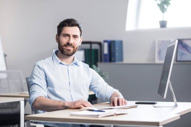 Beyaz gömlekli, bilgisayar masasında oturan yakışıklı bir gencin portresi. Kameraya bakıyor, gülümsüyor, çalışıyor.