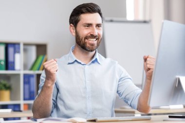 Genç, başarılı bir adam, ofis çalışanı, yönetici, serbest çalışan yapılan işin sonucuna seviniyor. Masada ve bilgisayarda mutlu bir şekilde oturur, sevinir, ellerini kaldırır.
