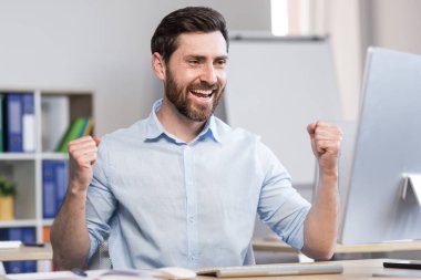 Genç, başarılı bir adam, ofis çalışanı, yönetici, serbest çalışan yapılan işin sonucuna seviniyor. Masada ve bilgisayarda mutlu bir şekilde oturur, sevinir, ellerini kaldırır.