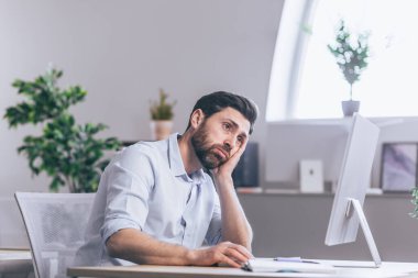 Tired and thoughtful office worker. Sitting at a desk in the office, at the computer, holding his head in his hands, tired, in a white shirt