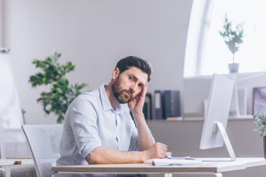 Yorgun ve düşünceli bir ofis çalışanı. Ofisteki bir masada oturuyor, bilgisayarın başında, başını elinde tutuyor, yorgun, beyaz bir gömlekle.