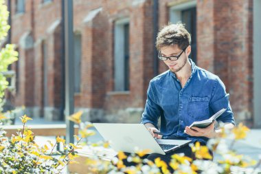 Kampüsün dışında genç bir öğrenci bankta ders çalışıyor. Bilgisayarlı bir adam internetten uzaktan çalışıyor.