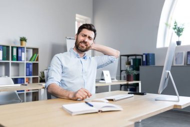 Bilgisayarda çalışan yorgun genç bir adam ofiste bir masada oturuyor boynundaki acıyı hissediyor, boynuna masaj yapıyor, rahatlıyor..
