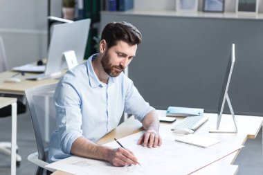 Genç bir mimar, tasarımcı, çizimler yapıyor ve ofisteki masanın üstündeki kağıt üzerinde proje yapıyor. Bilgisayarla bir masada oturup, odaklanmak...