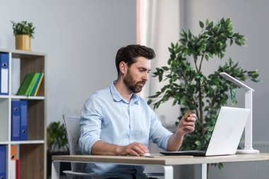 Resepsiyondaki bilgisayarda çalışan serbest çalışan genç bir adam. Kredi kartı tutuyor, hesapta çalışıyor, para kazanıyor, emirler yağdırıyor.