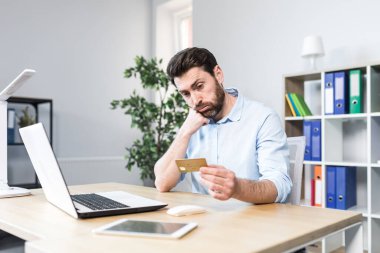 Başarısız çevrimiçi alım. Başarısız bir anlaşma. Üzgün genç bir adam, elinde kredi kartı olan bir ofis çalışanı, şok içinde başını tutuyor. Masada oturuyor, dizüstü bilgisayarın başında.