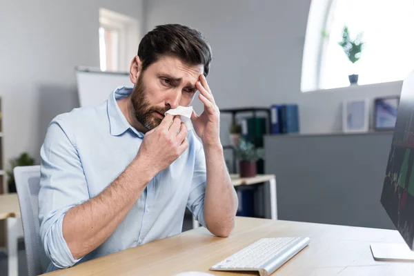 Sick and tired young man at work. Office worker, manager, freelancer sitting at a desk in the office does not feel well, wipes his nose, has a runny nose, coughs, headaches, holds his head.