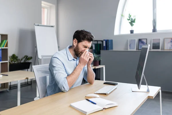 Sick and tired young man at work. Office worker, manager, freelancer sitting at a desk in the office does not feel well, wipes his nose, has a runny nose, coughs, headaches, holds his head.