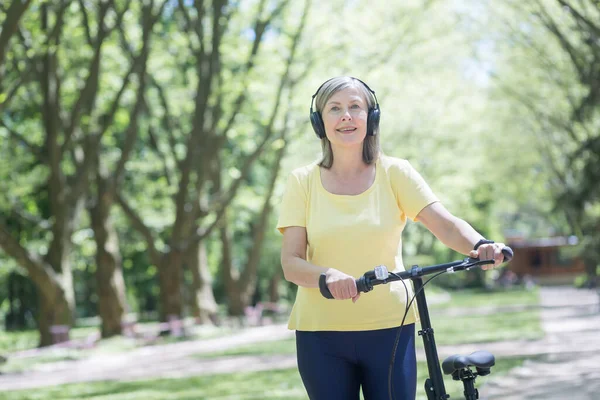 Senior happy woman on a walk in the park with a bicycle listens to music on headphones, and walks on a summer day