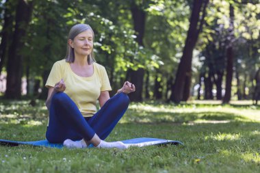Yaşlı, kır saçlı, emekli bir kadın parkta meditasyon yapıyor ve nefes egzersizleri yapıyor, yaz günü Lotus pozisyonunda.
