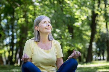Yaşlı, kır saçlı, emekli bir kadın parkta meditasyon yapıyor ve nefes egzersizleri yapıyor, yaz günü Lotus pozisyonunda.