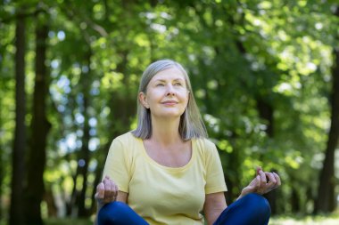 Yaşlı, kır saçlı, emekli bir kadın parkta meditasyon yapıyor ve nefes egzersizleri yapıyor, yaz günü Lotus pozisyonunda.