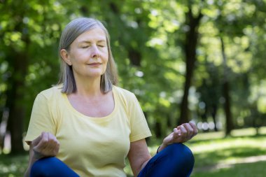 Yaşlı, kır saçlı, emekli bir kadın parkta meditasyon yapıyor ve nefes egzersizleri yapıyor, yaz günü Lotus pozisyonunda.