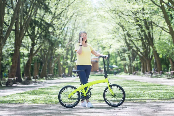 上了年纪的快乐女人在夏天的公园里散步 微笑着 用自行车在电话里聊天 — 图库照片