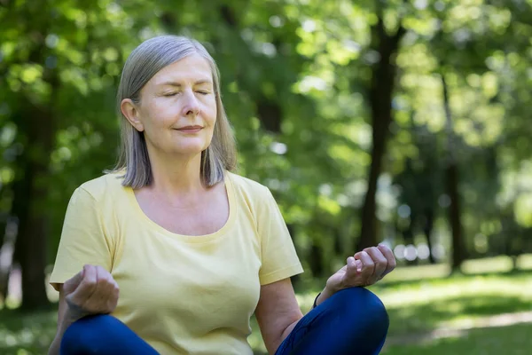Yaşlı, kır saçlı, emekli bir kadın parkta meditasyon yapıyor ve nefes egzersizleri yapıyor, yaz günü Lotus pozisyonunda.