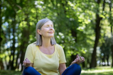 Aktif ve sağlıklı bir yaşam tarzı. Güzel, son sınıf bir kadının portresi. Yoga yapıyor. Parkta çimlerin üzerinde bir spor minderinin üzerinde oturur, gözlerini kapatır, dinlenir, meditasyon yapar.