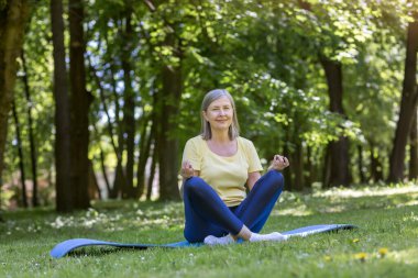 Aktif ve sağlıklı bir yaşam tarzı. Güzel, son sınıf bir kadının portresi. Yoga yapıyor. Parkta çimlerin üzerinde bir spor minderinin üzerinde oturur, dinlenir, meditasyon yapar.