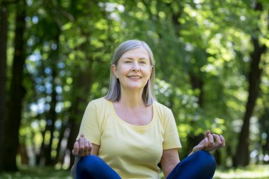 Aktif ve sağlıklı bir yaşam tarzı. Güzel, son sınıf bir kadının portresi. Yoga yapıyor. Parkta çimlerin üzerinde bir spor minderinin üzerinde oturuyor, kameraya bakıyor, gülümsüyor, dinleniyor, meditasyon yapıyor.