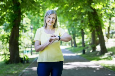 Aktif yaşam tarzı. Kıdemli güzel kadın sabah parkta koşuyor. Elindeki saatin nabzını ölçer ve görüntüler. Spor için gidiyor. İyi hissettiriyor, gülümsüyor.