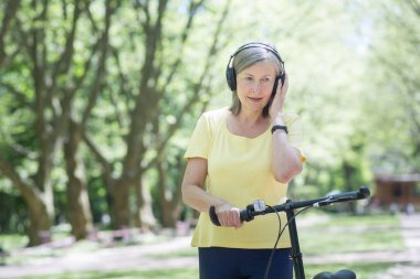 Aktif yaşam tarzı. Son sınıf güzeli kadın parkta yürüyor, sarı bir bisiklete biniyor, kulaklıkla müzik dinliyor. Elinde bir bisikletle duruyor, gülümsüyor, iyi vakit geçiriyor.