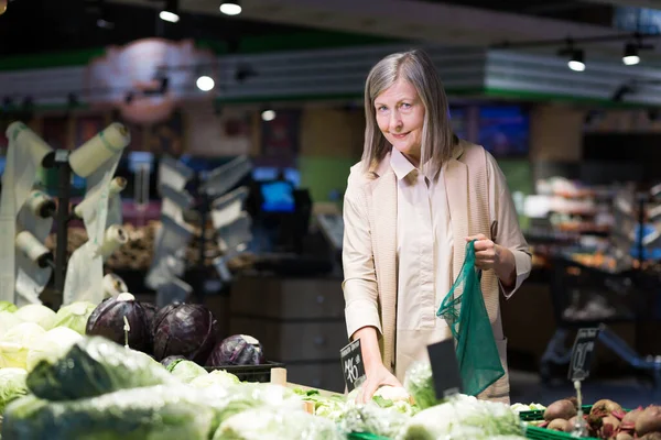 Portrait of a senior woman in a grocery supermarket, digs vegetables and puts in an ecological bag smiling and looking at the camera
