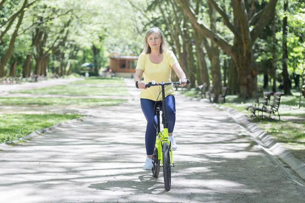 积极的生活方式 老漂亮的女运动员在公园里骑着一辆黄色的自行车 在户外 他走了 看着相机 — 图库照片