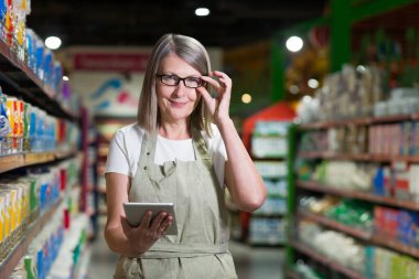 Gözlük danışmanı kıdemli kadın portresi, bakkal, süpermarket çalışanı. Tabletle duruyor, malları kabul ediyor, çekleri alıyor. İş kıyafetleriyle.