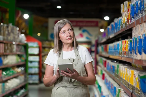 Portrait of senior female consultant, employee in grocery store, supermarket. Stands with a tablet, accepts the goods, checks. In work clothes.