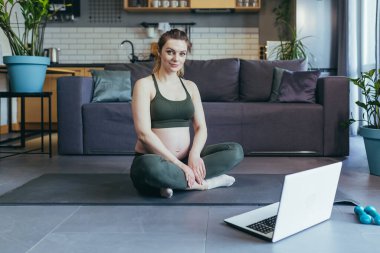 Evde dizüstü bilgisayardan spor yapan hamile güzel bir kadın spor yapıyor, spor minderinin üzerinde oturuyor, egzersiz yapıyor, mutlu.