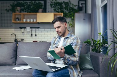 Uzaktan öğrenme çevrimiçi. Genç adam, öğrenci. Evde dizüstü bilgisayarla, kitaplarla çalışıyor. Kanepede oturmak bir deftere çevrimiçi ders yazar. Dinler ve dersleri izler.