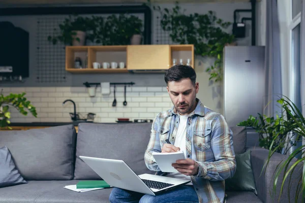 Uzaktan öğrenme çevrimiçi. Genç adam, öğrenci. Evde dizüstü bilgisayarla, kitaplarla çalışıyor. Kanepede oturmak bir deftere çevrimiçi ders yazar. Dinler ve dersleri izler.