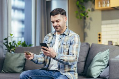 Kanepede oturan bir adam telefonu online alışveriş için kullanıyor, elinde bir banka kartı var.