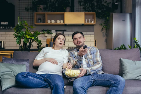 Home cinema. Young family. Pregnant woman and man sitting on sofa at home, watching TV, movie, eating popcorn. Have a good time together