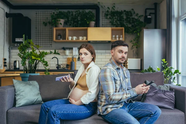 Ignore. Young couple, pregnant woman and man sitting on sofa at home with their backs to each other, holding mobile phones in their hands. Do not talk to each other. They look at the camera, disappointed