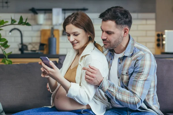 Young family sitting on sofa at home. Pregnant woman and man. Woman holding a phone in her hands shows her husband, calling together parents, friends. A man hugs a woman from behind. They are smiling