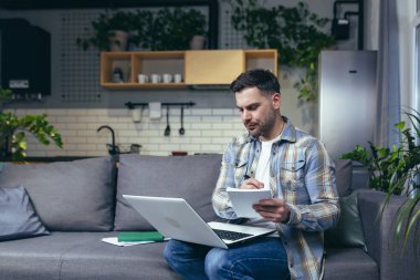 Evdeki adam kanepede dizüstü bilgisayarla oturuyor.