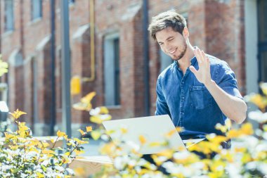 Sakallı genç bir adam video görüşmesi için dizüstü bilgisayar kullanıyor, soğuk bir adam gülümsüyor ve bilgisayar ekranına uzaktan bakan arkadaşlarıyla konuşuyor.