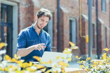 Sakallı genç bir adam video görüşmesi için dizüstü bilgisayar kullanıyor, soğuk bir adam gülümsüyor ve bilgisayar ekranına uzaktan bakan arkadaşlarıyla konuşuyor.