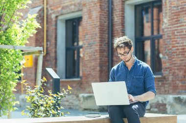 Kampüsteki genç öğrenci laptopta çalışıyor ve çalışıyor, ofis dışında günlük kıyafetler giymiş bir adam.