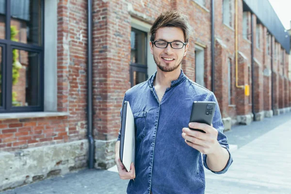 Kampüsün dışında elinde cep telefonuyla gülümseyen ve yürüyen genç bir öğrencinin portresi.