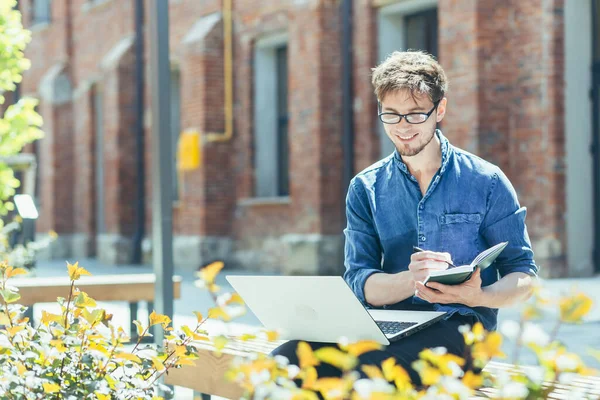 Üniversite kampüsünün dışındaki genç bir öğretmenin portresi dizüstü bilgisayarla bankta oturuyor ve gülümsüyor.