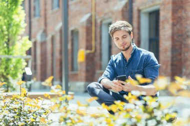Genç bir adam, bir adam, bir öğrenci güneşli bir havada ofis merkezinin yanındaki bankta oturuyor, elinde cep telefonu kullanıyor..