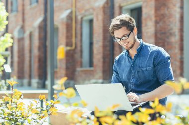 Genç adam, stajyer öğrenci. İnternette bir dizüstü bilgisayarda çalışıyor, bir proje üzerinde çalışıyor, ofis merkezinin yanındaki bankta oturuyor, gözlük takıyor..