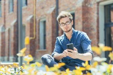 Genç bir adam, bir adam, gözlüklü ve kot pantolonlu bir öğrenci güneşli bir havada ofis merkezinin yanındaki bankta oturuyor, cep telefonu kullanıyor, elinde tutuyor. s