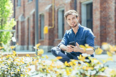 Genç bir adam, bir adam, güneşli bir havada ofis merkezinin yanındaki bankta oturan bir öğrenci, elinde cep telefonu kullanıyor. Kameraya bakıyor, gülümsüyor.