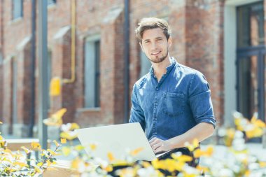Genç ve yakışıklı bir erkek öğrencinin portresi. İnternette bir dizüstü bilgisayarda çalışıyor, ofis merkezinin yanındaki bankta oturuyor. Kameraya bakıyor, gülümsüyor.